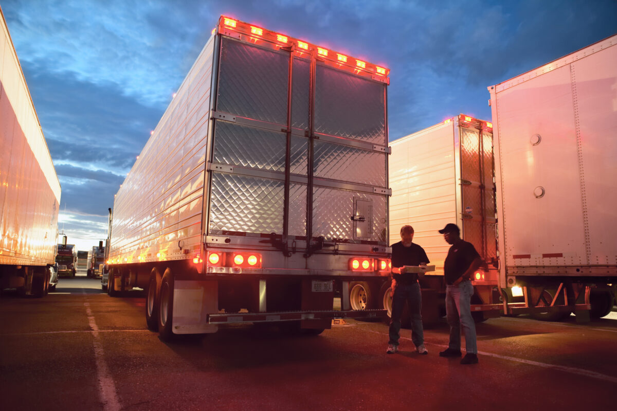 Two truck drives checking dispatch papers while standing next to truck trailers in a large parking lot at night. Freight Brokerage
